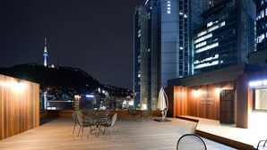 Une terrasse sur le toit, dotée d’un plancher en bois, de chaises et de tables en métal. On aperçoit des bâtiments en arrière-plan, avec des fenêtres illuminées.