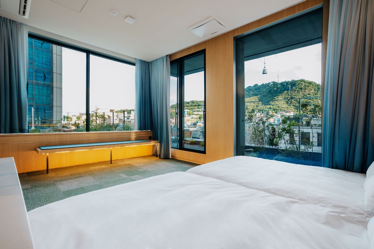 A hotel room with a large bed, a bench, and a view of a cityscape through large windows.