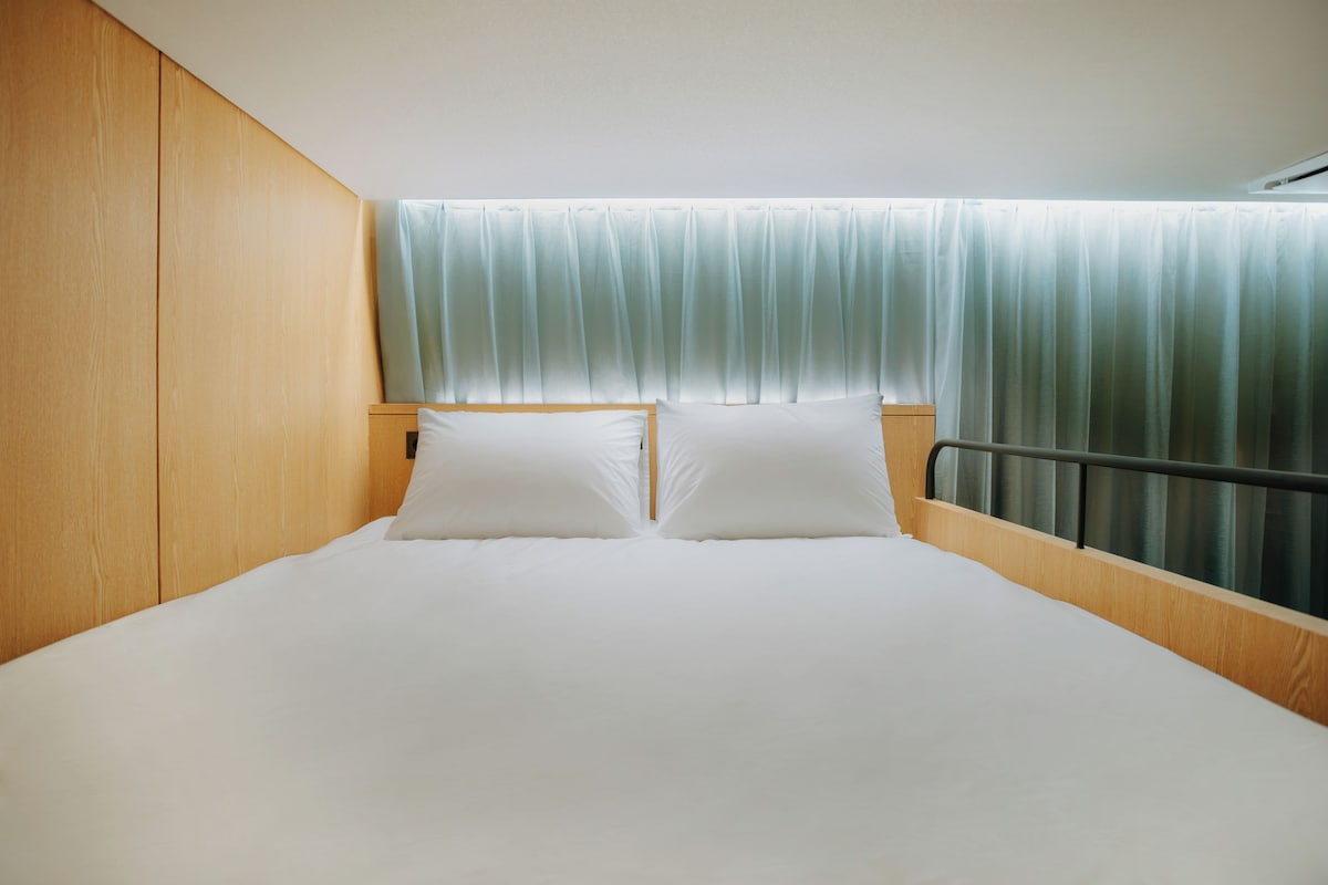 A single bed with a wooden headboard and a window with sheer curtains.
