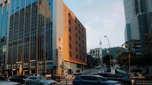 A multi-story hotel with a prominent vertical sign, a modern glass building adjacent to it, and a busy street with vehicles and pedestrians. - HOTEL DRIP&DROP Myeongdong Central (Seoul)