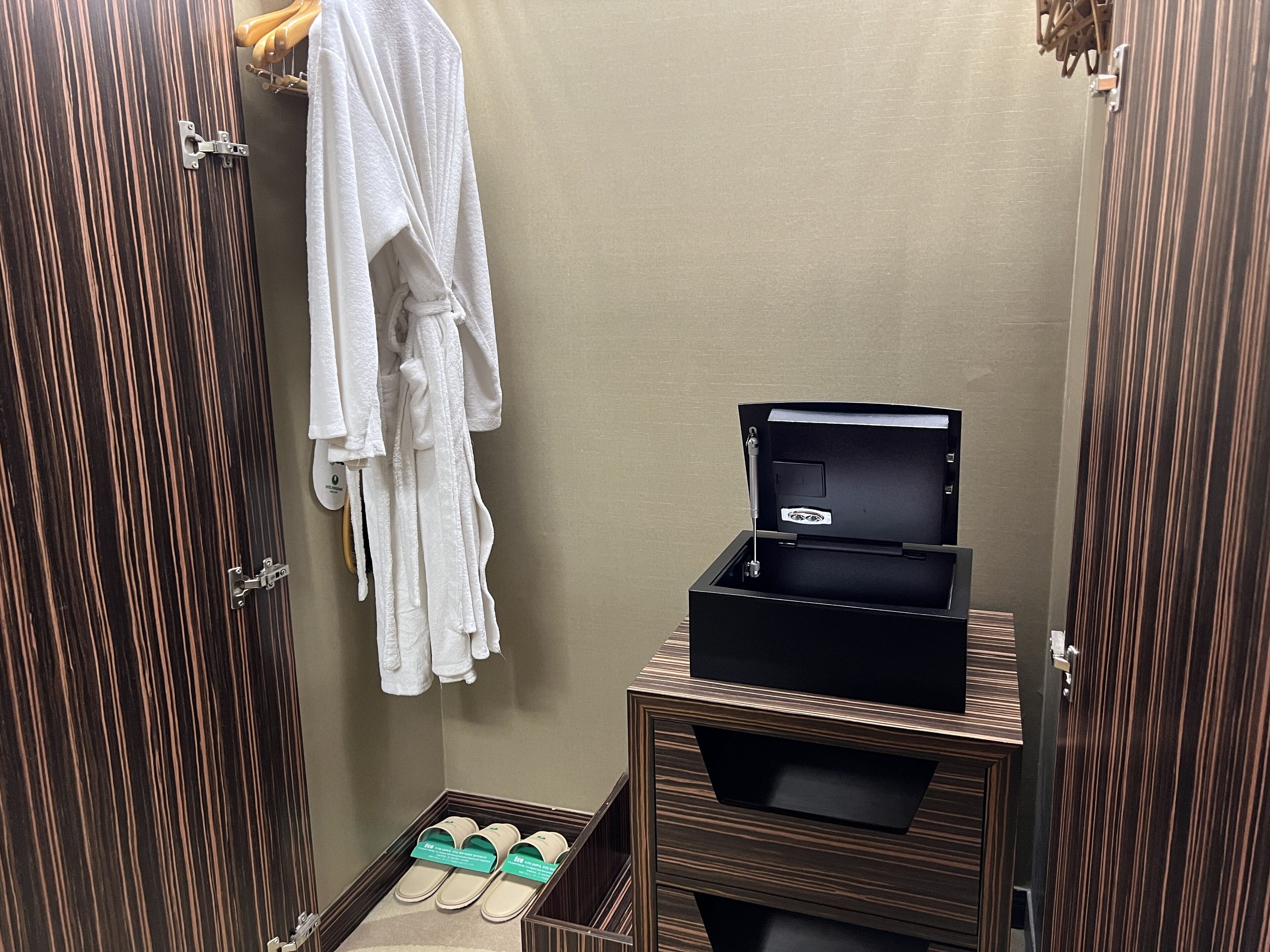 A small bathroom with a wooden vanity, a robe hanging on the wall, a black storage box, and a pair of slippers on the floor.