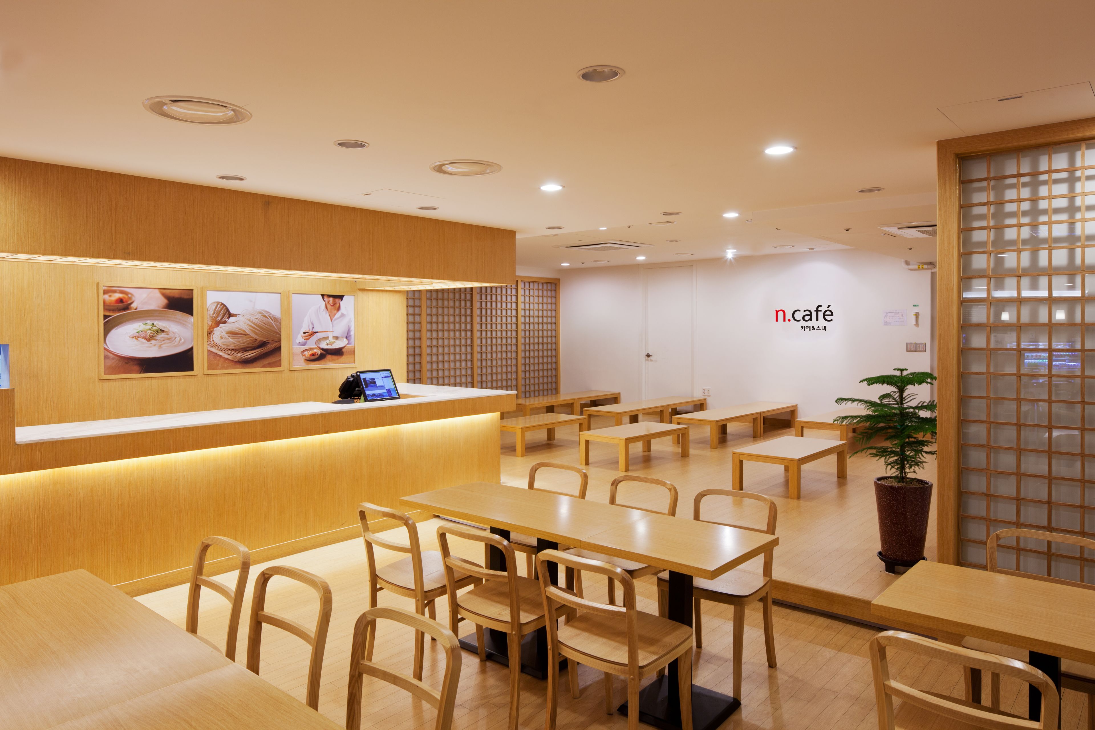 A cafe interior with wooden tables and chairs, a counter with a menu display, and a potted plant.