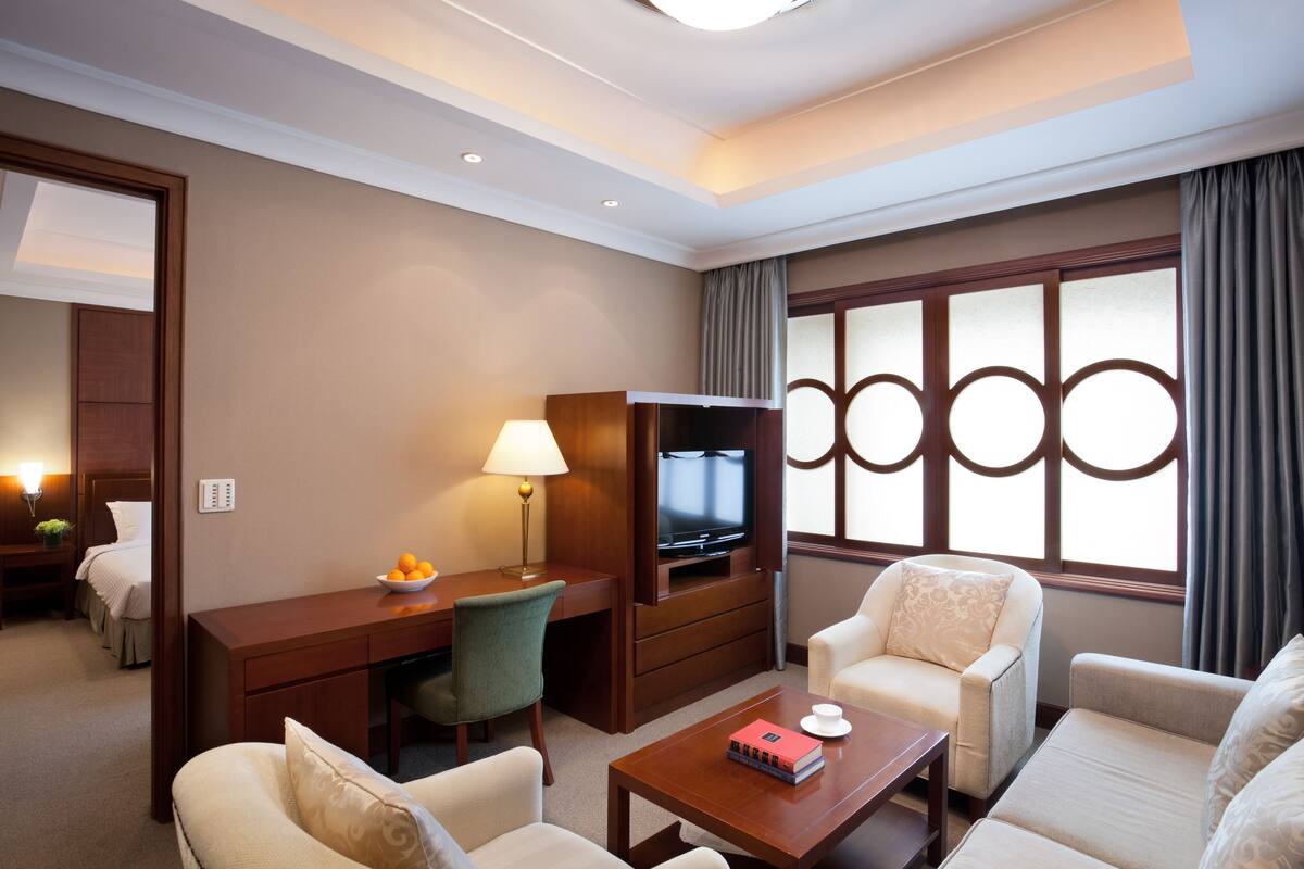 Habitación de hotel con escritorio, televisor, dos sillones, una mesa de centro y un ventanal con cortinas.