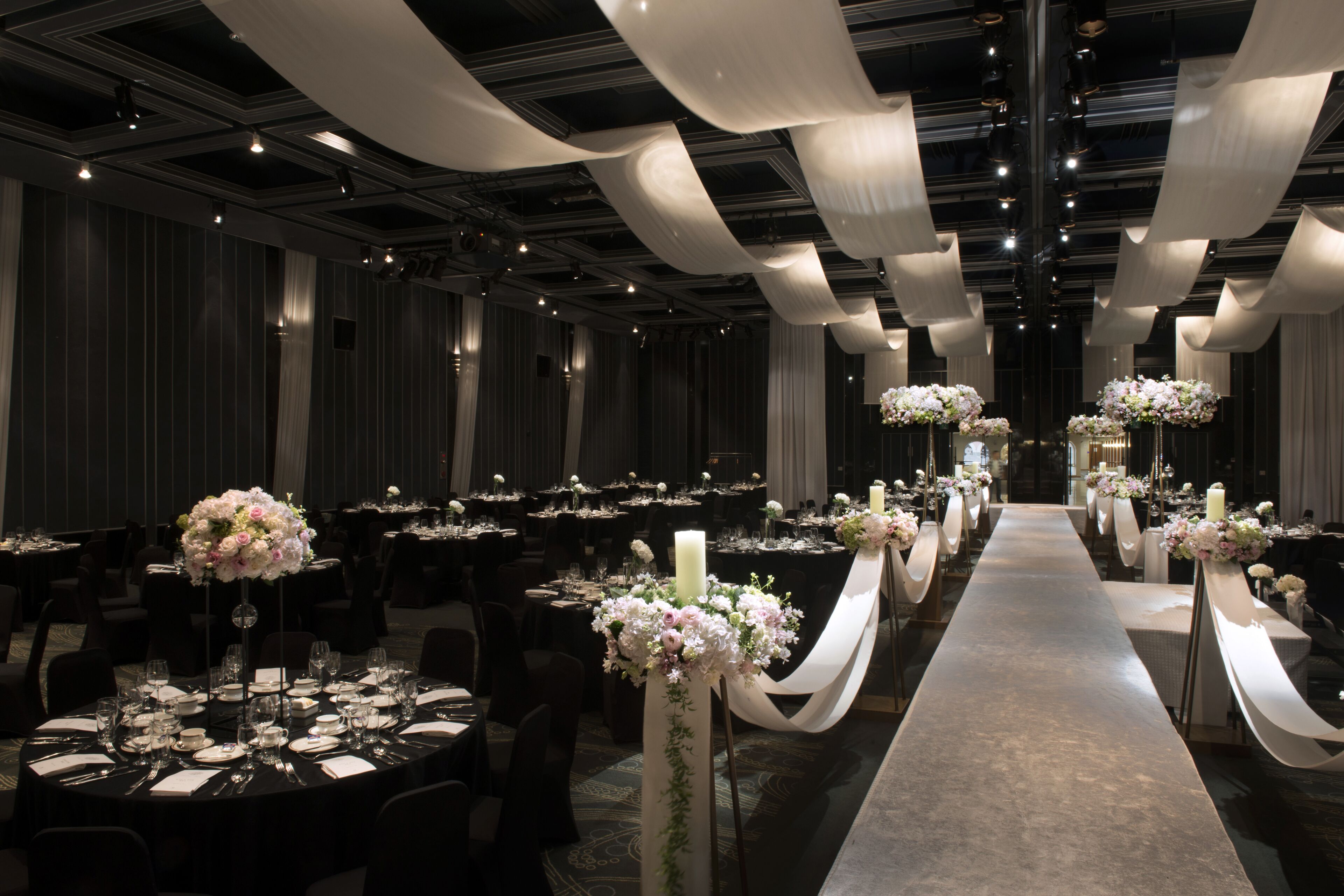 A banquet hall with round tables, white drapes, and floral centerpieces.