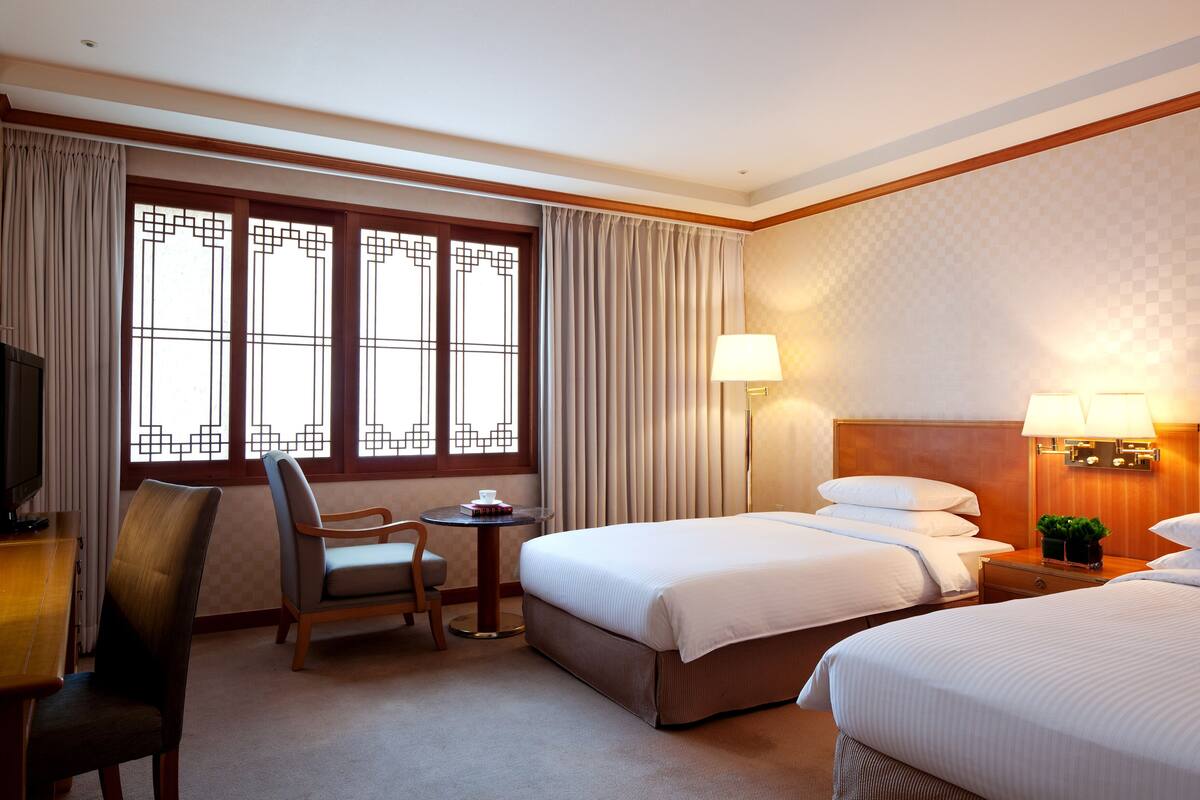 Una habitación de hotel con dos camas, un escritorio con silla, un televisor y una ventana con cortinas.