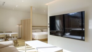 Et moderne hotelværelse med seng, stort vægmonteret TV, spiseplads med bord og stole samt et separat siddeområde med sofa og lille bord.