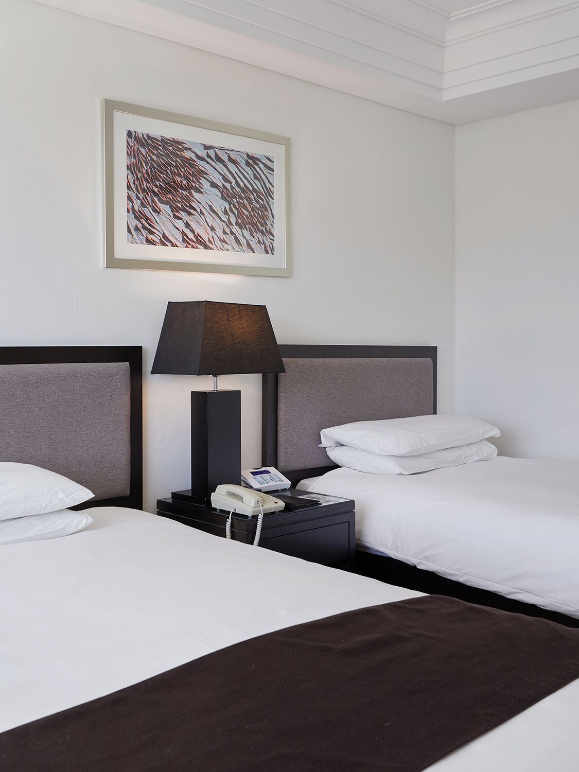 Habitación de hotel con dos camas, una mesita de noche con lámpara, teléfono y un cuadro abstracto enmarcado en la pared.