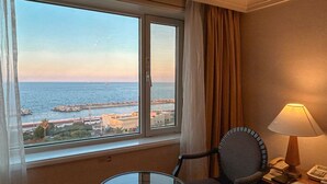 A hotel room with a large window offering a view of the sea and a distant cityscape. There is a round glass table with a chair, a bedside lamp, and a telephone on a bedside table. - Jeju Oriental Hotel & Casino (Jeju City)