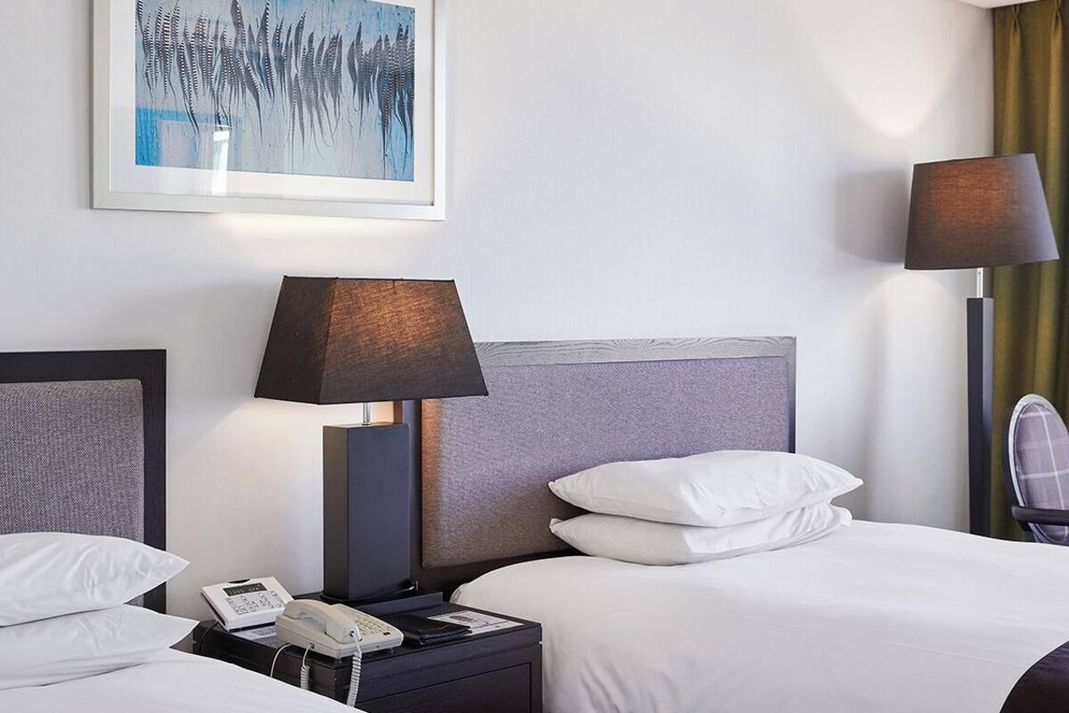 A hotel room with two beds, a bedside table with a lamp, a phone, and a painting on the wall.