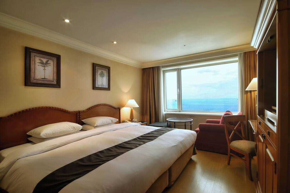 A hotel room with a large bed, a desk, a chair, a small round table, and a view of the sea through a large window.