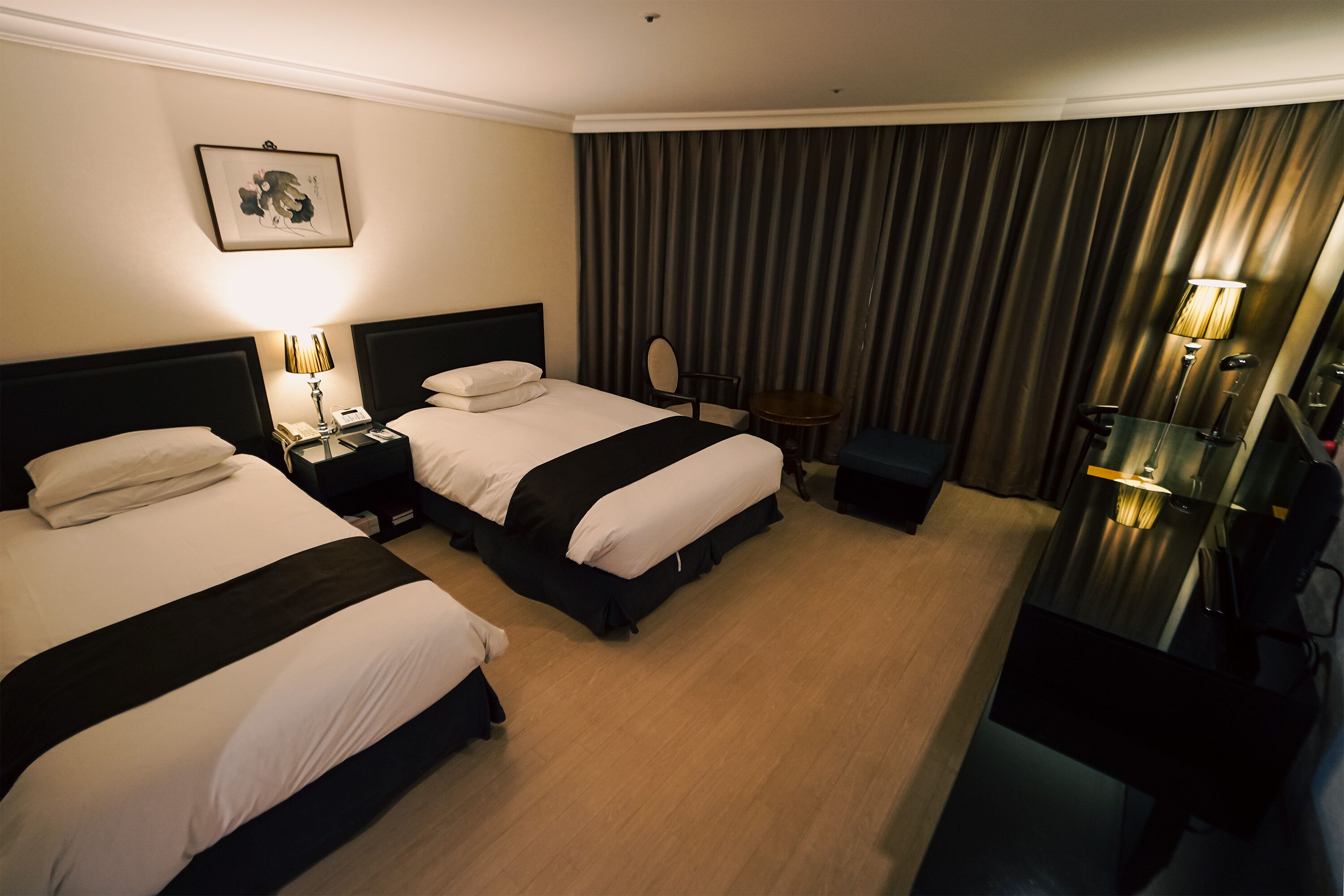Een hotelkamer met twee bedden, een bureau, een stoel, een tafeltje, een lamp en een gordijn.