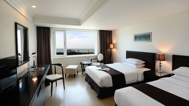 A hotel room with two beds, a desk, a chair, a TV, and a window with a view. - Jeju Oriental Hotel & Casino (Jeju City)