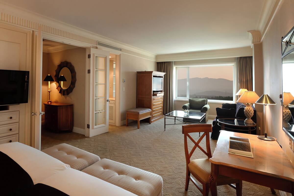 A hotel room with a bed, a desk, a TV, a chair, a lamp, a dresser, a mirror, and a view of the mountains.