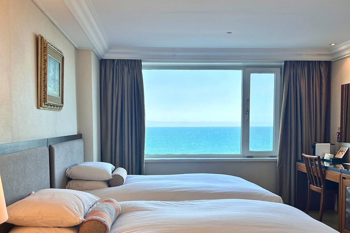 A hotel room with two beds, a desk, and a view of the sea.