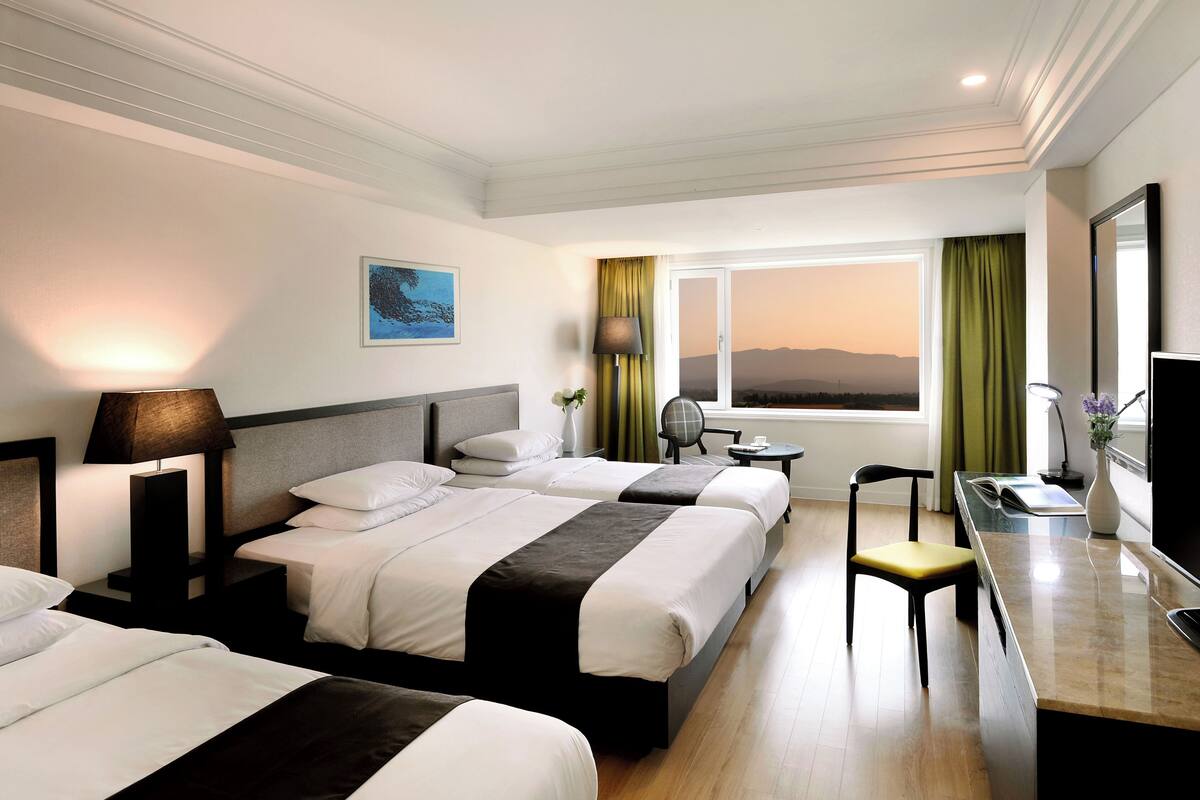 A hotel room with two beds, a desk, a chair, a TV, and a window with a view.