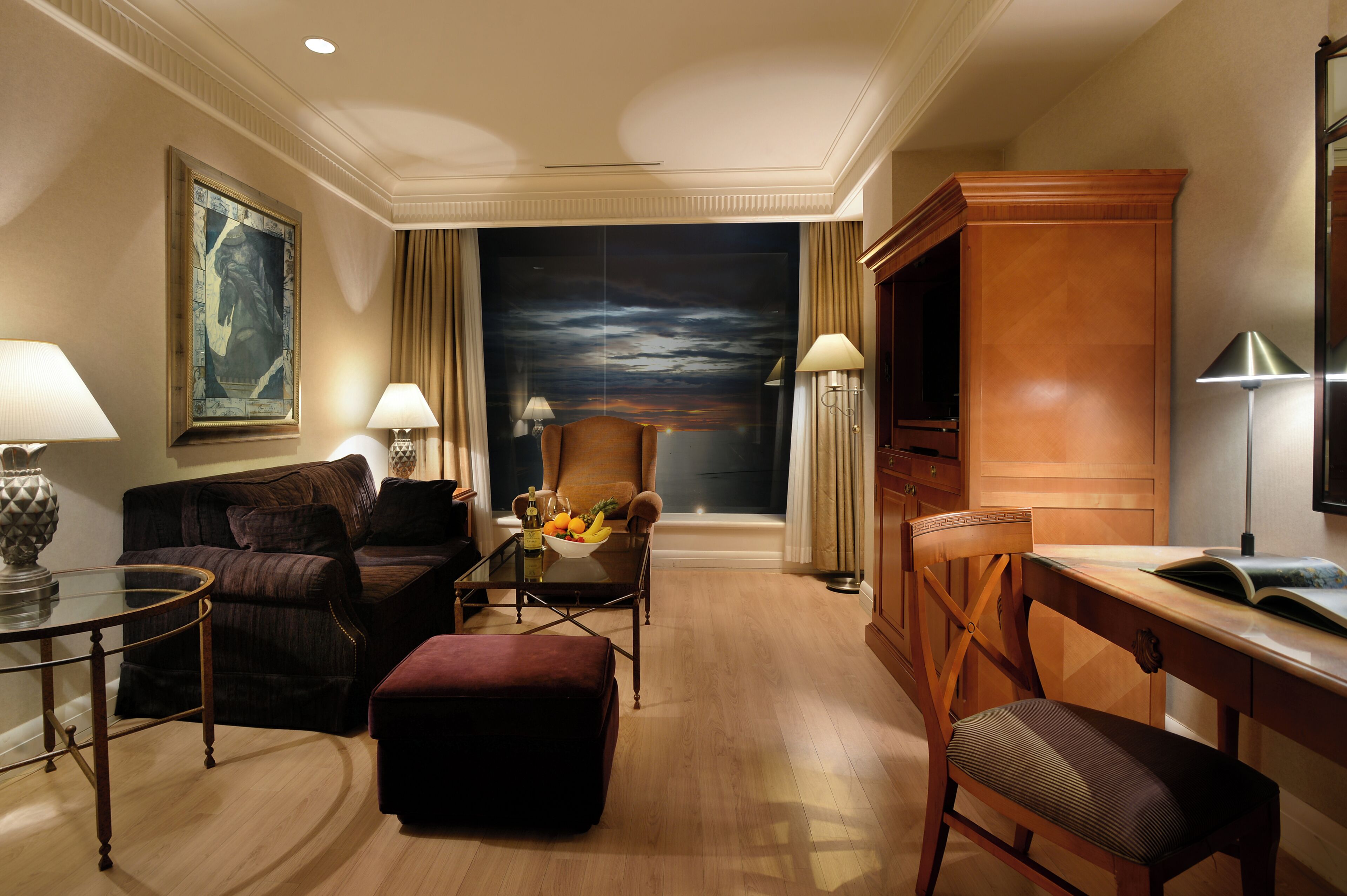 A hotel room with a large painting, a sofa, a chair, a desk, and a dining table with a fruit bowl.
