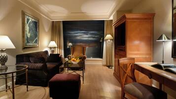 A hotel room with a large painting, a sofa, a chair, a desk, and a dining table with a fruit bowl.