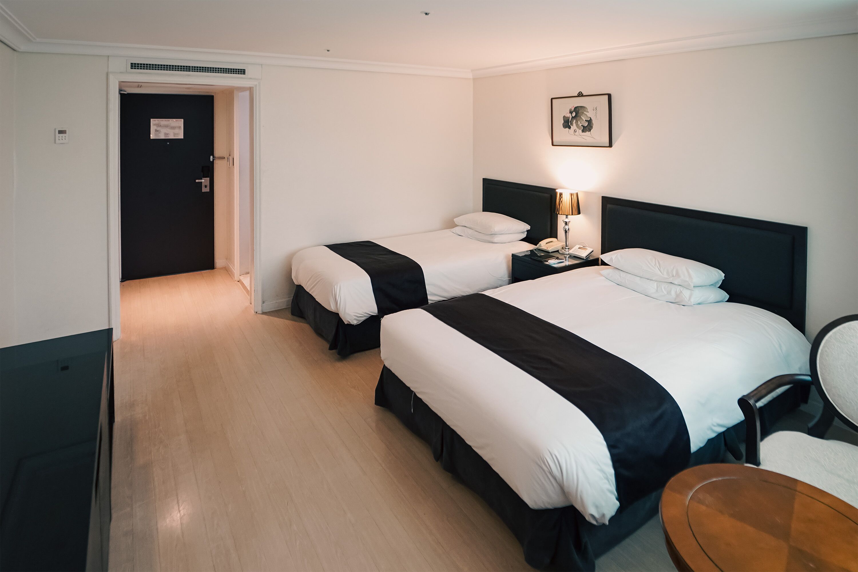 A hotel room with two beds, a bedside table with a lamp, a chair, and a wooden table.