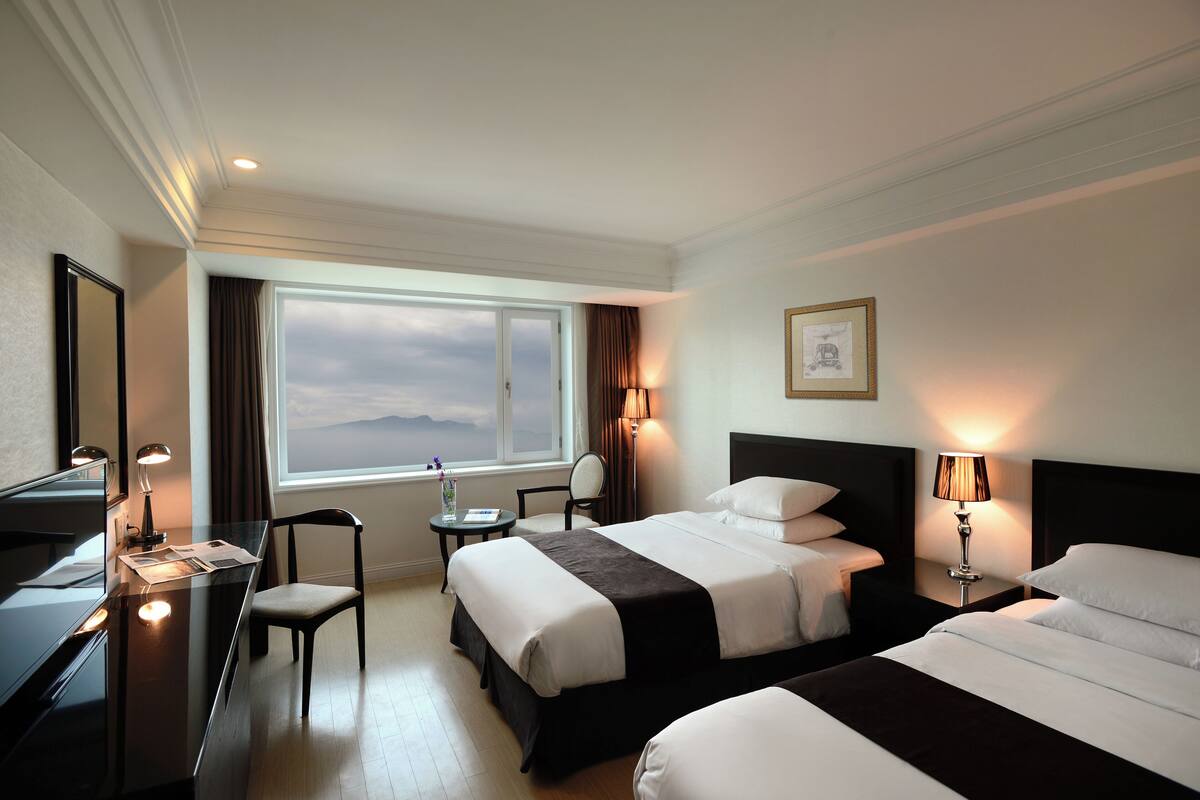 A hotel room with two beds, a desk, a chair, a window with a view, and a framed picture on the wall.