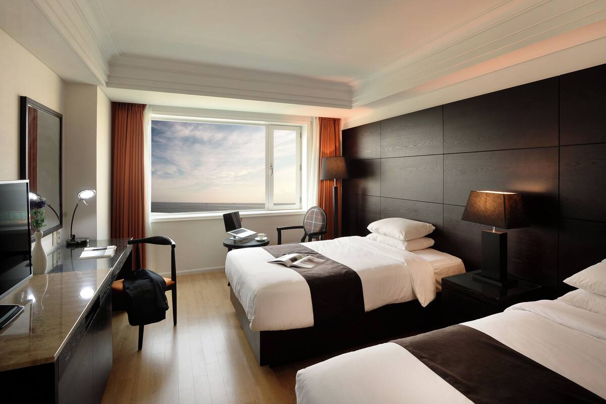 A hotel room with two beds, a desk with a computer, a TV, and a window with curtains.