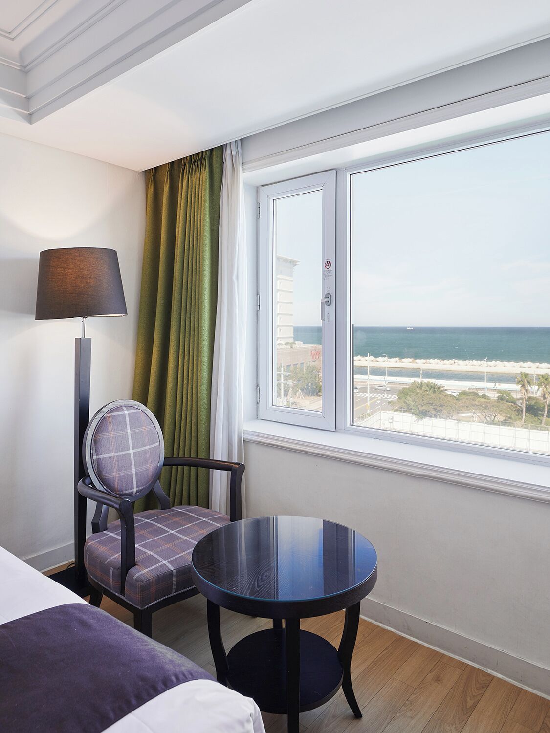 A hotel room with a bed, a chair, a table, and a large window offering a view of the beach and ocean.