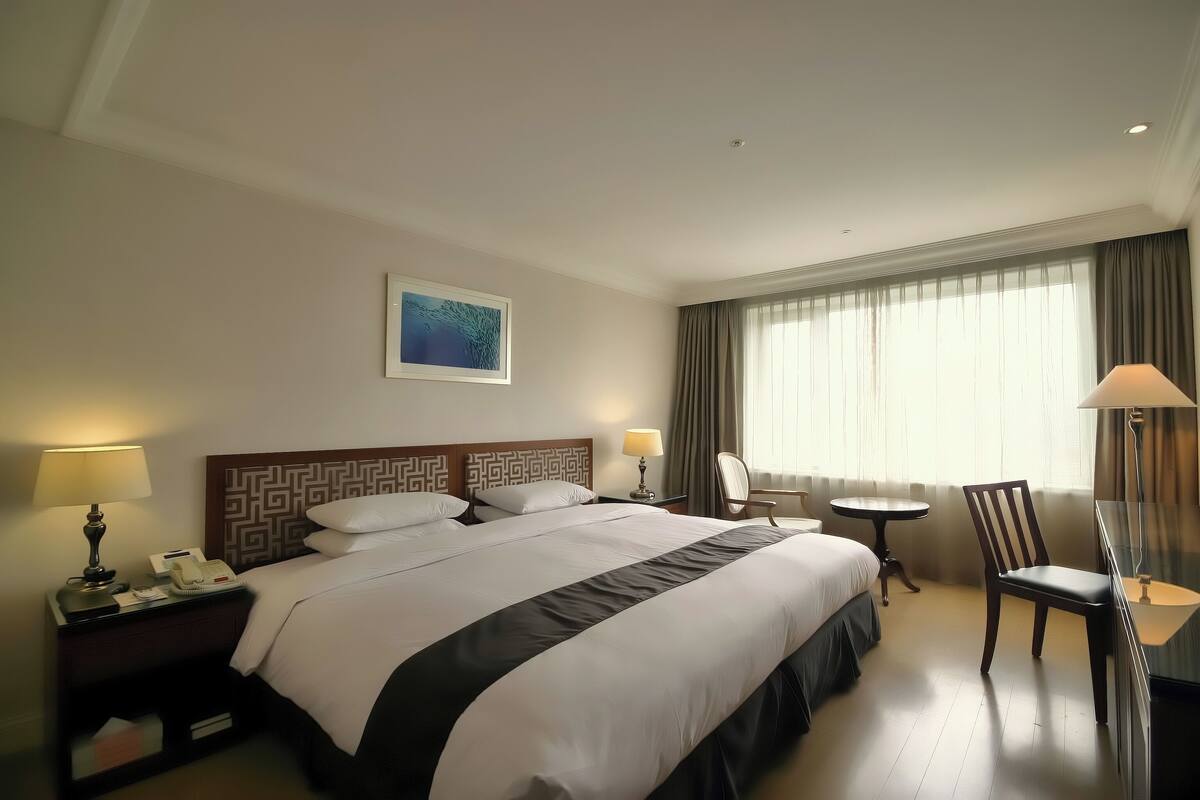 A hotel room with a large bed, two bedside tables with lamps, a chair, a small round table, and a large window with curtains.