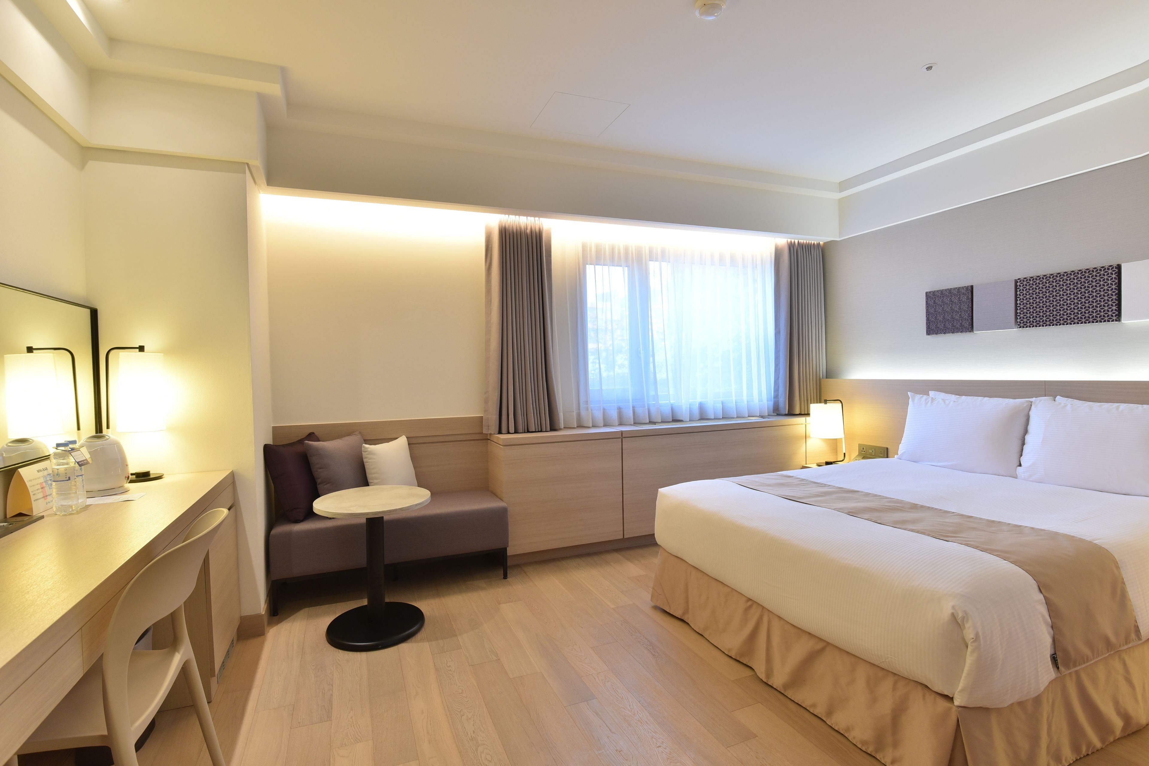 A hotel room with a bed, a desk, a chair, a small table, a sofa, and a window with curtains.
