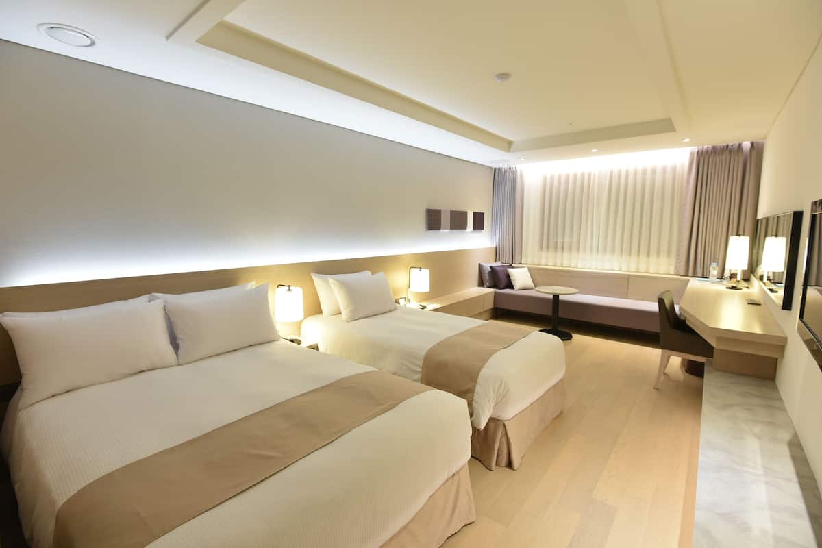 A hotel room with two beds, a sofa, a desk, and a large window with curtains.