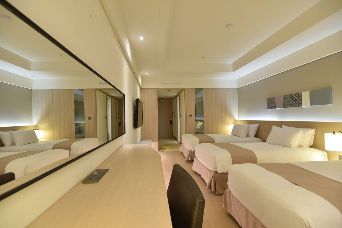 A hotel room with four beds arranged in a row, a large mirror, a desk with a chair, and a wall-mounted TV.