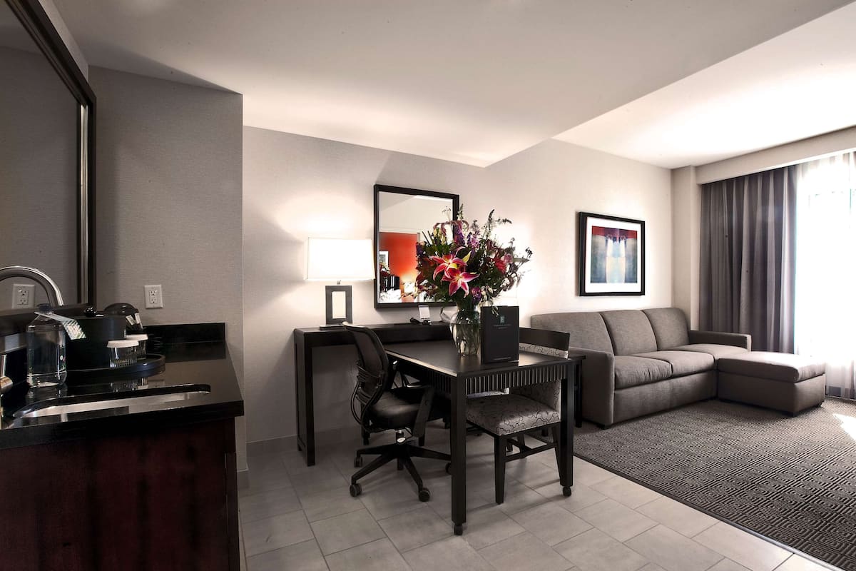 A modern hotel room with a dining area, a sofa, a desk with a chair, and a large mirror.