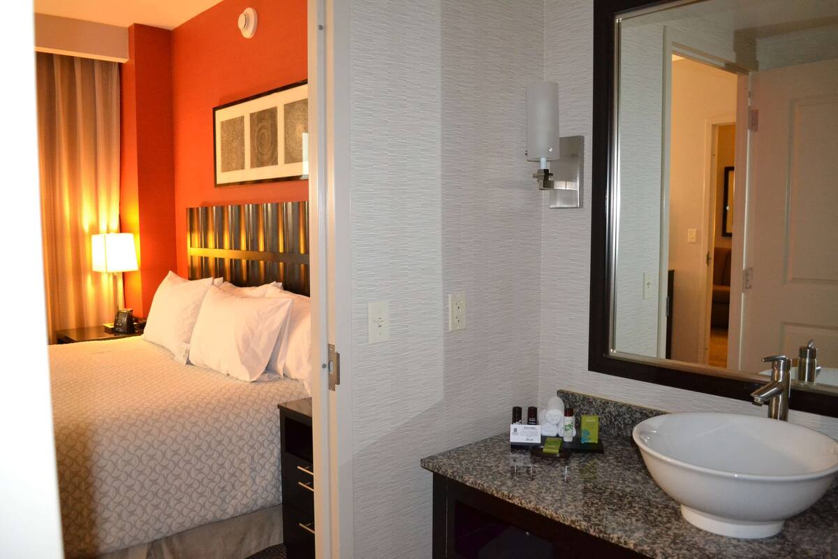 A hotel room with a bed, a nightstand, a lamp, a large mirror, and a bathroom sink.