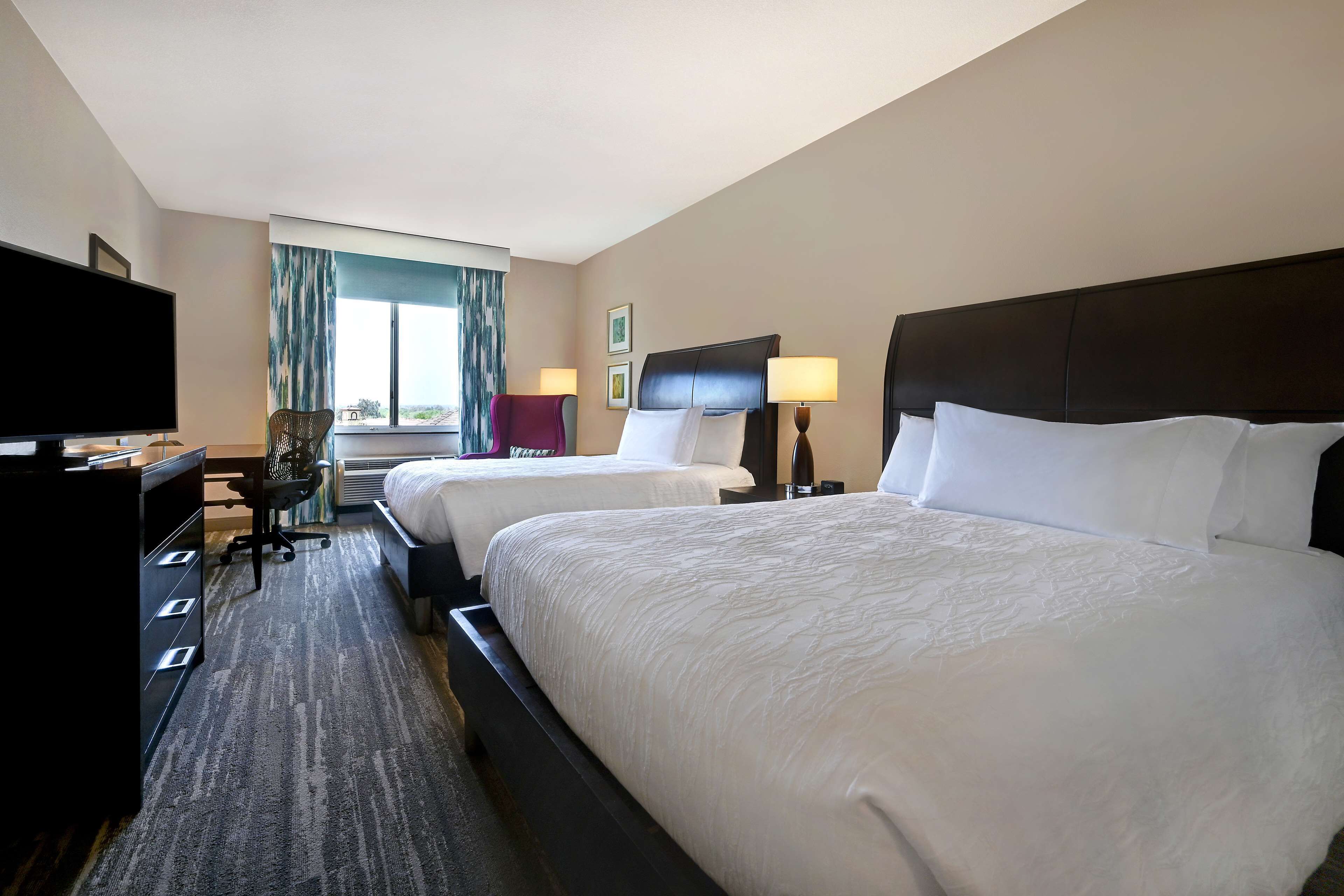Quarto de hotel com duas camas, uma escrivaninha, televisão de tela plana, uma cadeira e uma janela com cortinas.