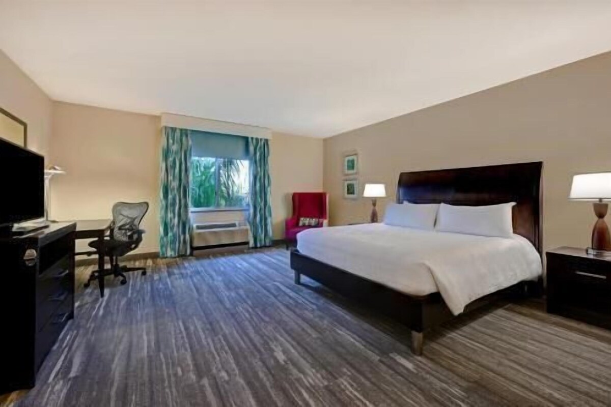 Habitación de hotel con cama, escritorio, silla, televisión y ventana con cortinas.