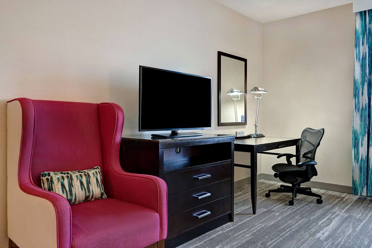 A hotel room with a red armchair, a desk with a computer, a flat-screen TV, and a mirror.