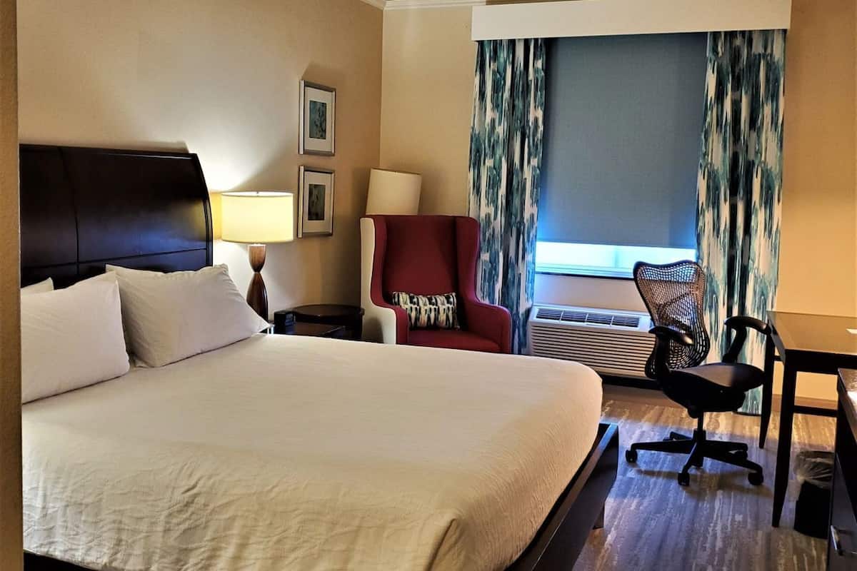 A hotel room with a large bed, a desk, a chair, a lamp, and a window with curtains.