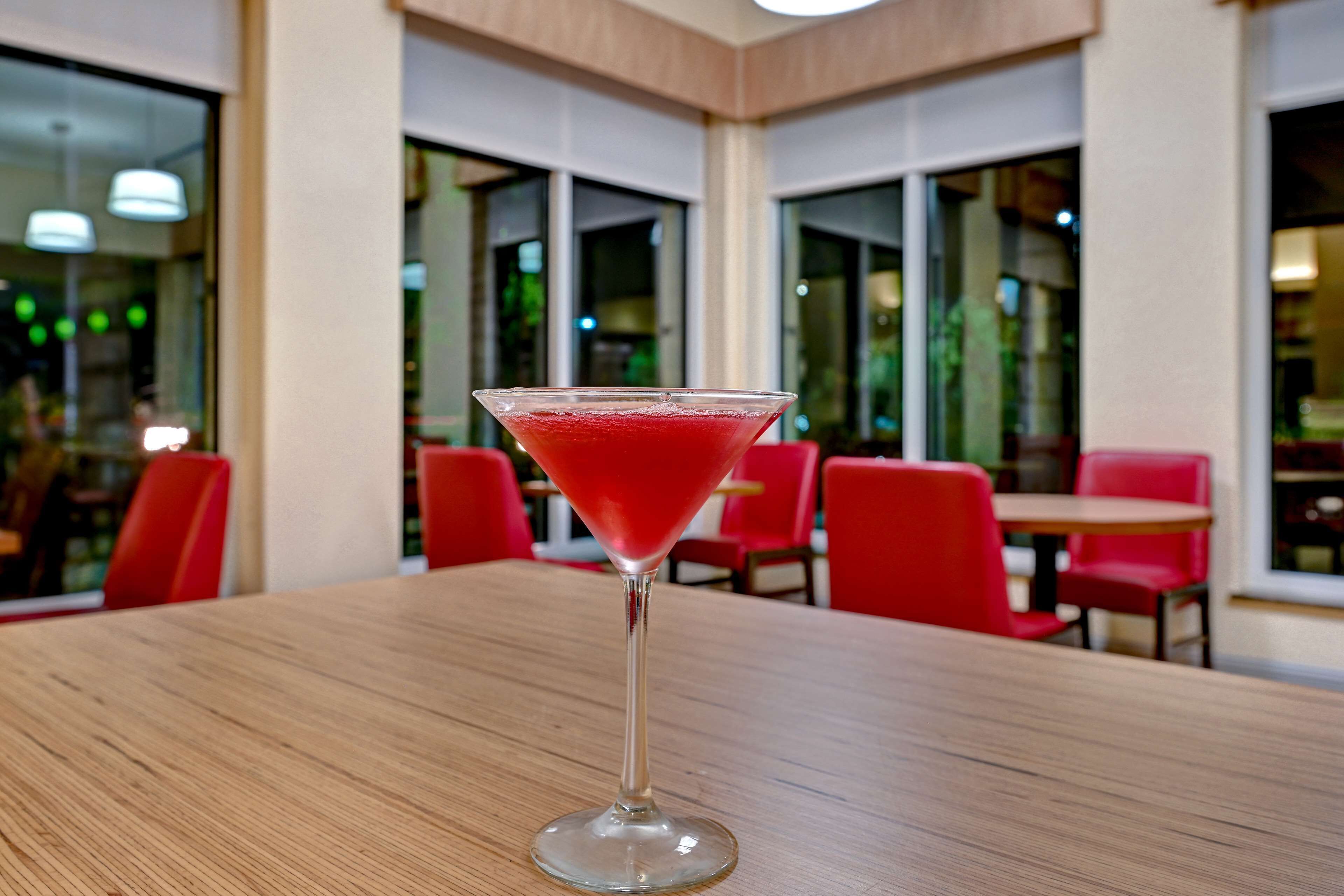 A martini glass with a red drink on a wooden table in a restaurant with red chairs and large windows.