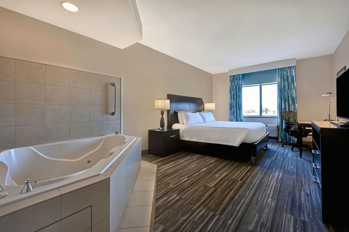 Habitación de hotel con una bañera grande, una cama con dos almohadas, un escritorio con una silla y una ventana con cortinas.