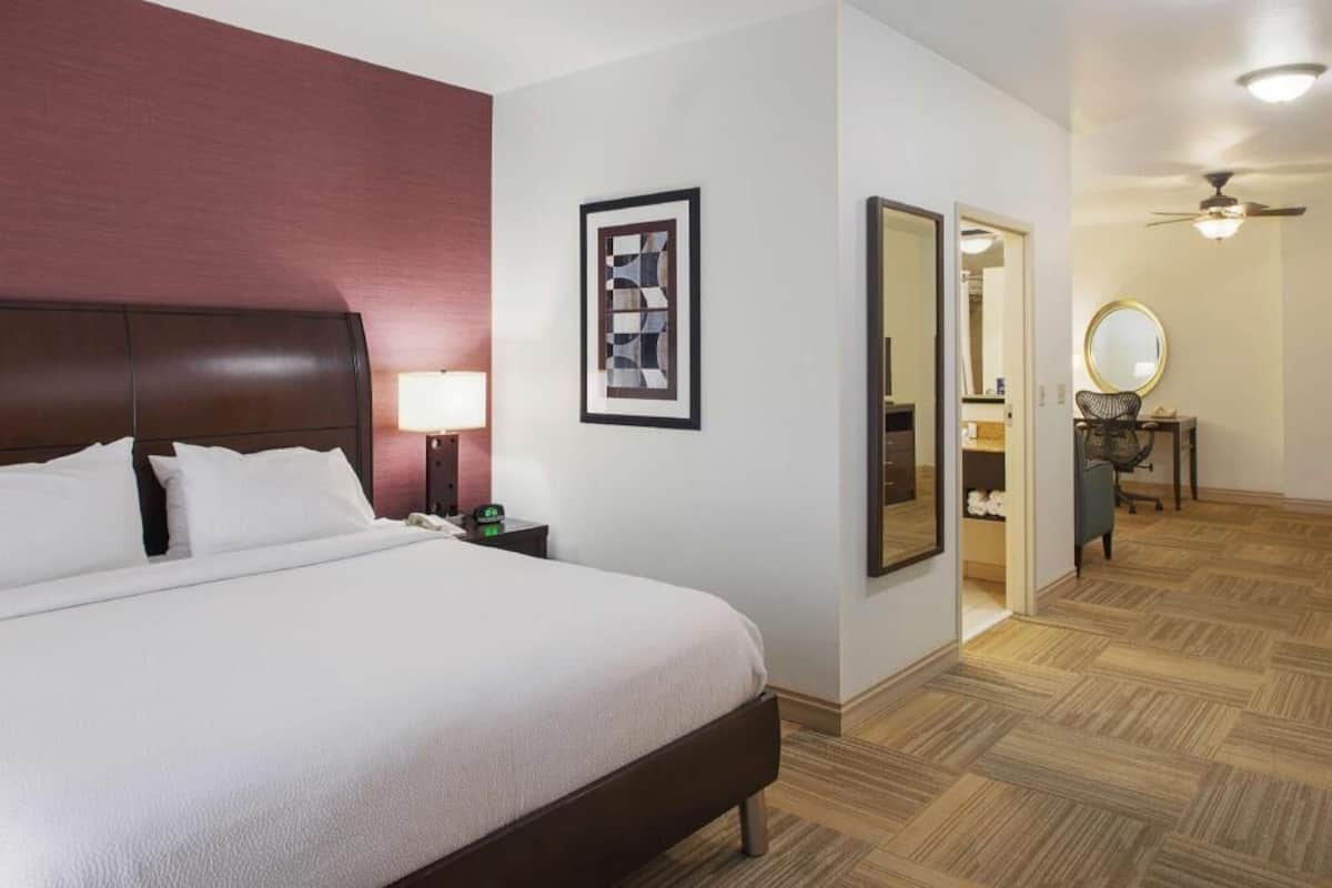 A hotel room with a large bed, a bedside table with a lamp, a framed abstract artwork, a mirror, and a ceiling fan.