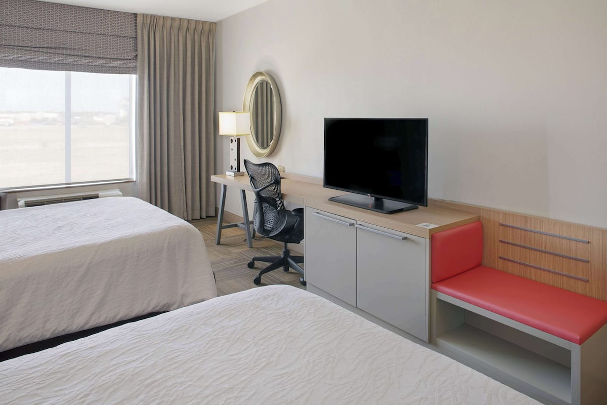 A hotel room with two beds, a desk with a chair, a television, and a small seating area with a red bench.