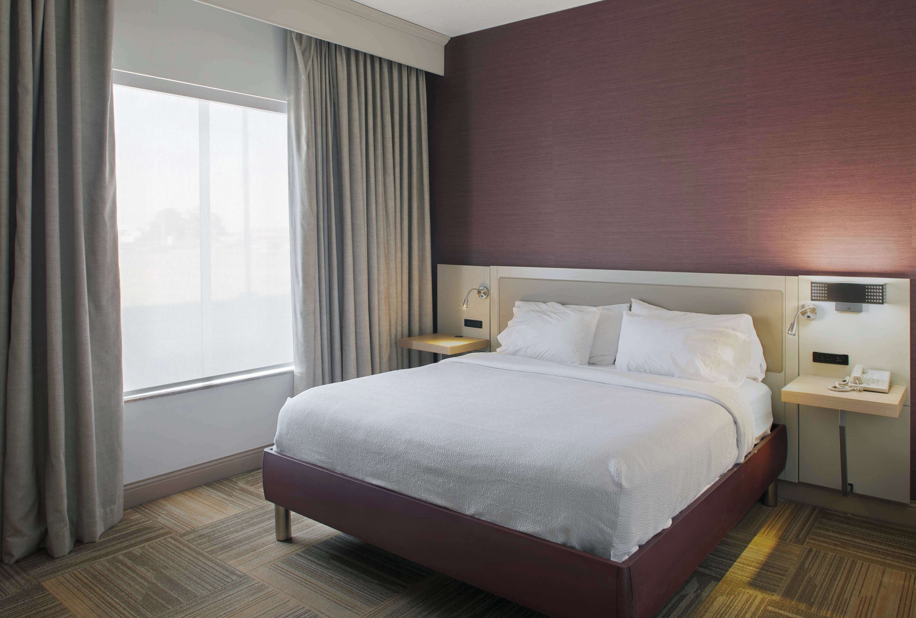 Una habitación de hotel con una cama grande, una mesita de noche y un ventanal con cortinas.