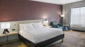 Hotelkamer met een groot bed, nachtkastje met lamp, stoel, klein tafeltje en raam met gordijnen.