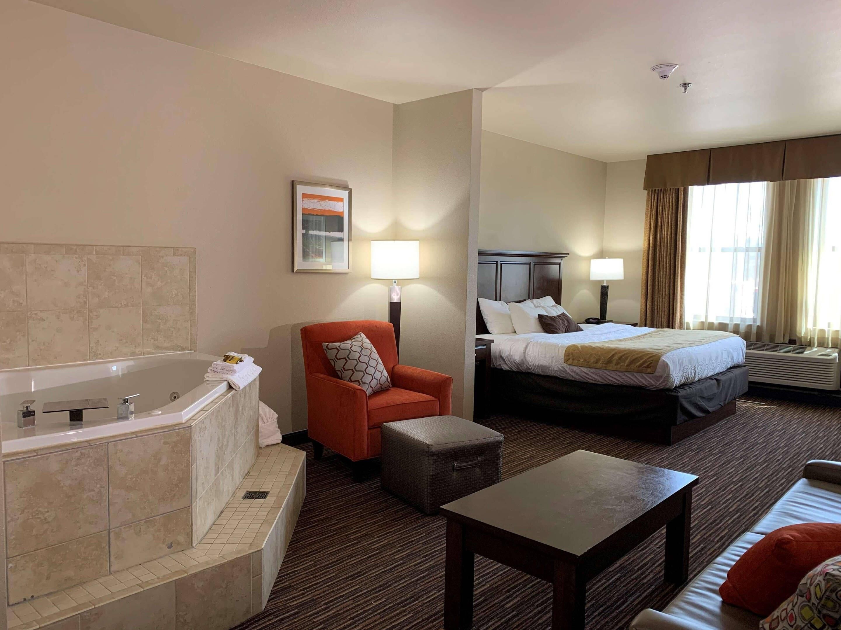 A hotel room with a bathtub, a bed with two pillows, a chair, a table, and a large window with curtains. - Radisson Hotel River Falls (River Falls)