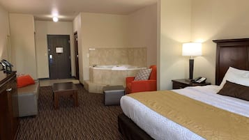 Une chambre d’hôtel comprenant un lit, un bureau, une chaise, une petite table, un canapé, une salle de bain avec baignoire, et une table de chevet avec une lampe et un téléphone.