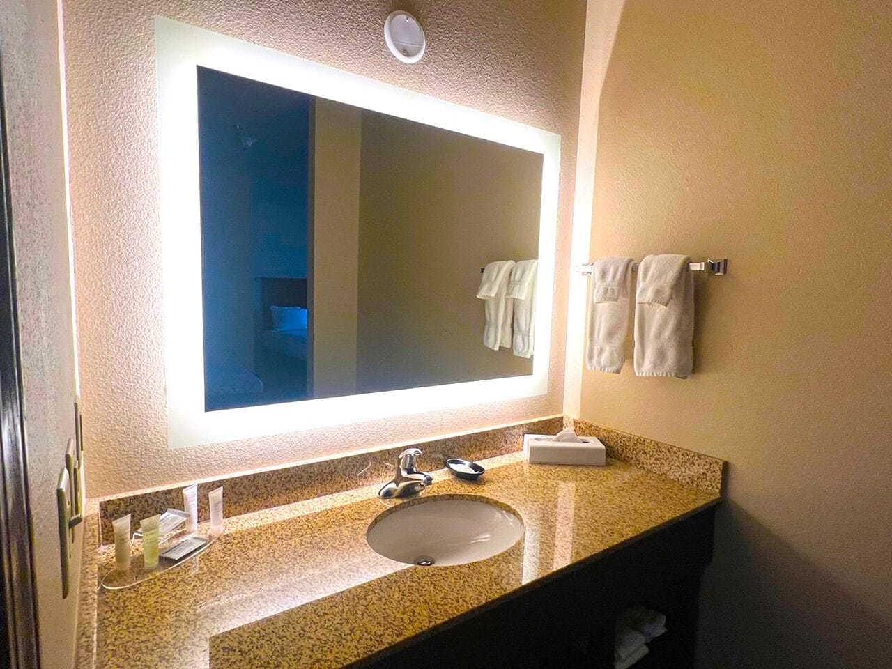 A bathroom with a granite countertop sink, a large mirror, and a towel rack with towels.