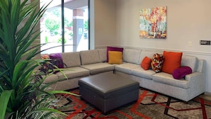 A modern living area with a sectional sofa, a grey ottoman, and a colorful abstract painting on the wall. - Radisson Hotel River Falls (River Falls)