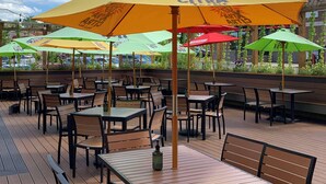 An outdoor patio with multiple tables and chairs, some under umbrellas with logos, and a view of a building in the background. - Radisson Hotel River Falls (River Falls)