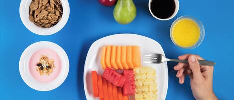 Une assiette contenant des carottes en tranches, de l’ananas et de la pastèque, un bol de céréales, une poire, une pomme et une tasse de café.