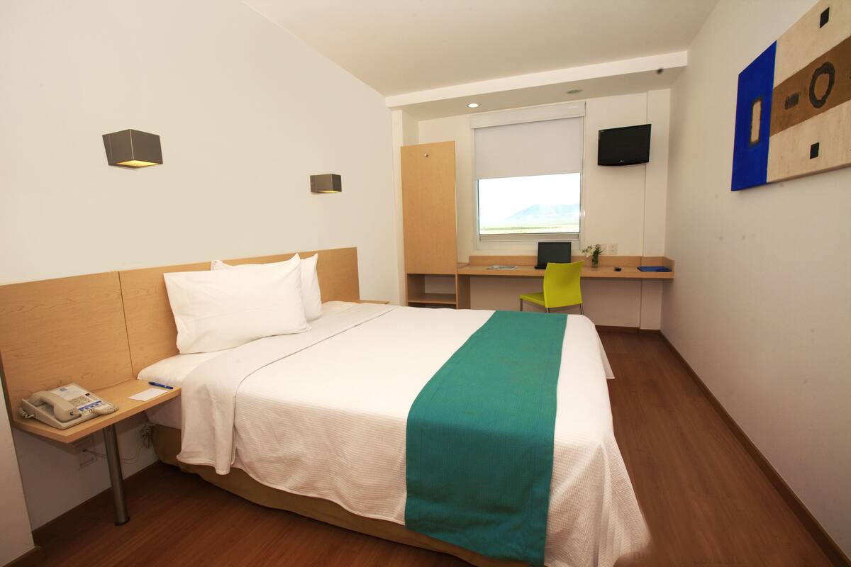 A hotel room with a bed, a desk with a computer, a chair, a phone, and a TV on the wall.