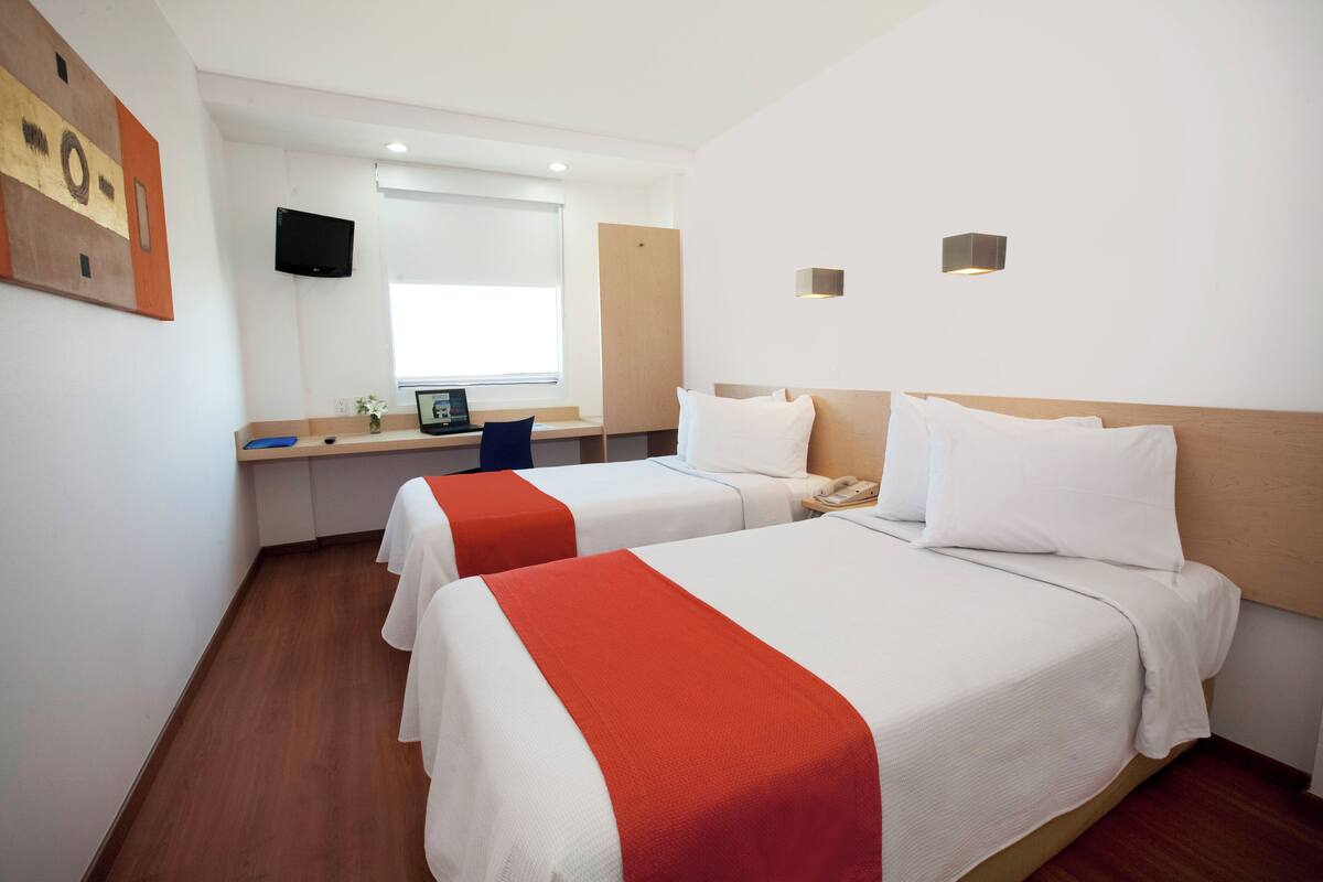 A hotel room with two beds, a desk with a computer, a TV, and a window.