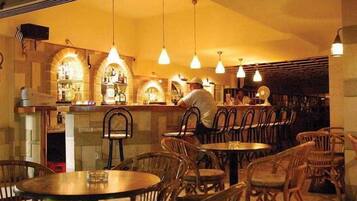 Interior de restaurante com mesas e cadeiras de madeira, luminárias pendentes e um bar com prateleiras cheias de garrafas.
